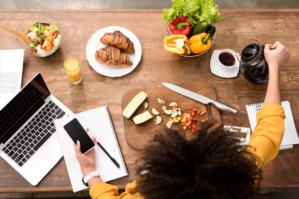 Vista superior del teléfono inteligente mujer joven en la cocina con varios alimentos y portátil en la mesa y verter café en la taza - foto de stock