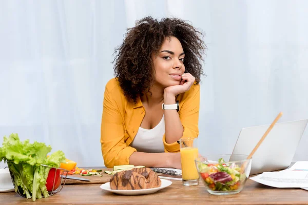 Jeune femme afro-américaine appuyée sur la table avec divers aliments et ordinateur portable, concept freelance — Photo de stock