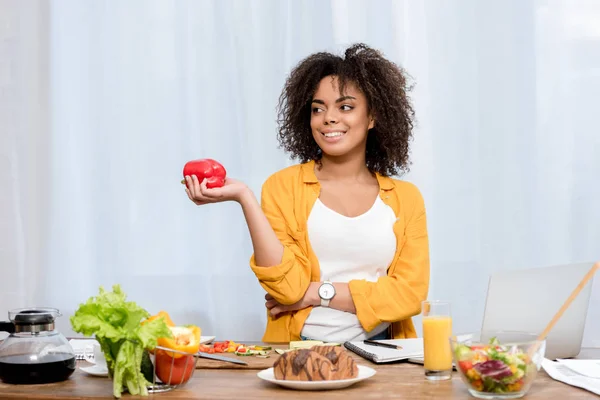 Schöne junge Frau mit verschiedenen Lebensmitteln und Arbeitsmaterialien auf dem Tisch, die zu Hause arbeitet — Stockfoto