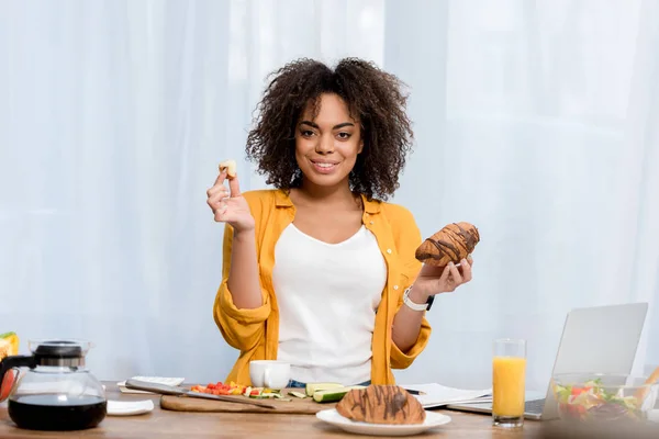 Schöne junge Frau isst Croissant, während sie zu Hause arbeitet — Stockfoto