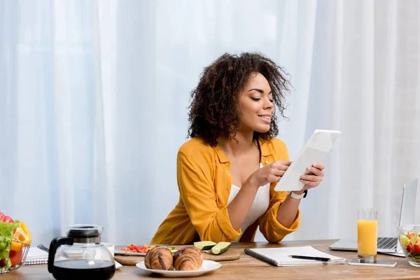 Mujer joven de raza mixta usando tableta en la cocina con comida en la mesa - foto de stock