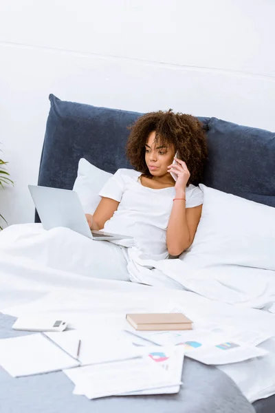 Jeune femme travaillant avec ordinateur portable et parlant par téléphone au lit à la maison — Photo de stock