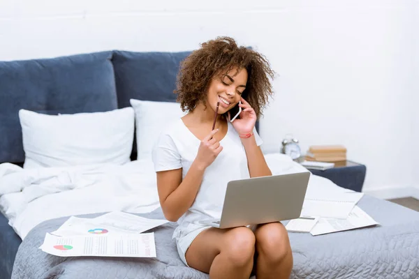 Щаслива молода жінка працює з ноутбуком і розмовляє по телефону на ліжку вдома — стокове фото