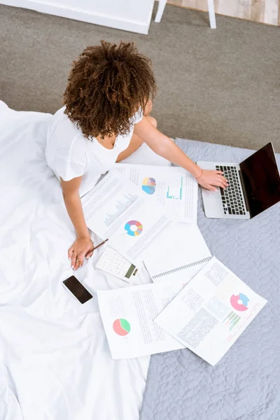 Vista de ángulo alto de la mujer que trabaja con el ordenador portátil y documentos en la cama en casa - foto de stock