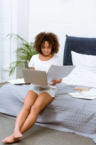 Bella giovane donna che lavora con computer portatile e documenti sul letto a casa — Foto stock