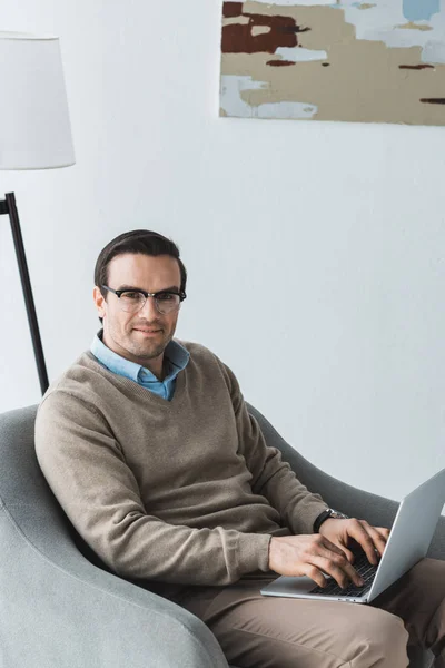 Homme dans des lunettes assis dans une chaise et travaillant sur un ordinateur portable — Photo de stock