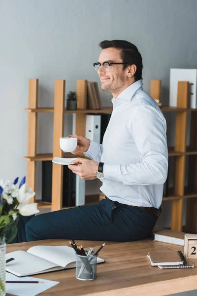 Uomo d'affari sorridente seduto sul tavolo e con in mano una tazza di caffè in un ufficio moderno — Foto stock