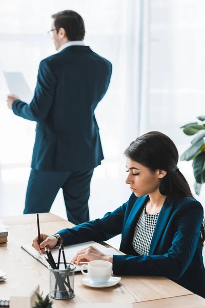 Imprenditrice che lavora al tavolo con taccuino mentre l'uomo studia carte alla finestra — Foto stock