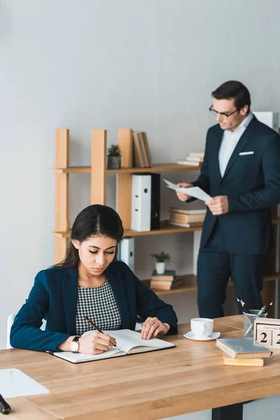Geschäftsfrau arbeitet am Tisch mit Notizbuch, während Mann im hellen Büro Papiere studiert — Stockfoto