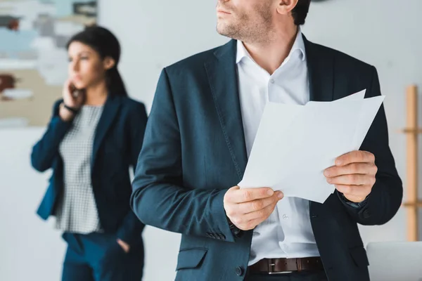 Geschäftsmann hält Papiere in der Hand, während Frau im hellen Büro telefoniert — Stockfoto