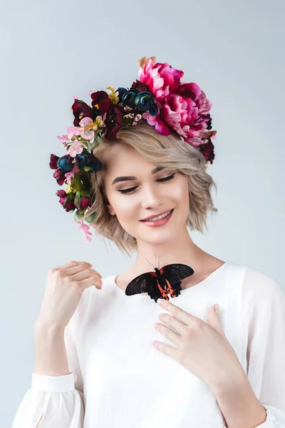 Усміхнена дівчина позує в квітковому вінку з живим метеликом на шиї, ізольована на сірому — стокове фото
