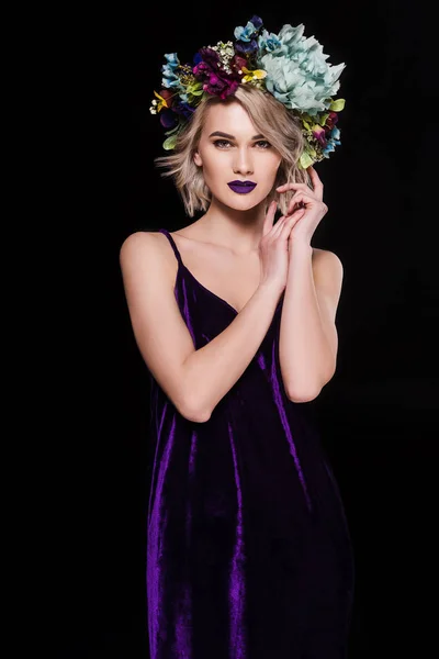 Красива молода жінка позує в фіолетовій сукні і квітковому вінку, ізольована на чорному — стокове фото