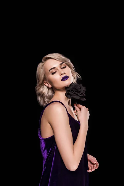 Chica rubia con los ojos cerrados posando en vestido púrpura con rosa negro, aislado en negro - foto de stock