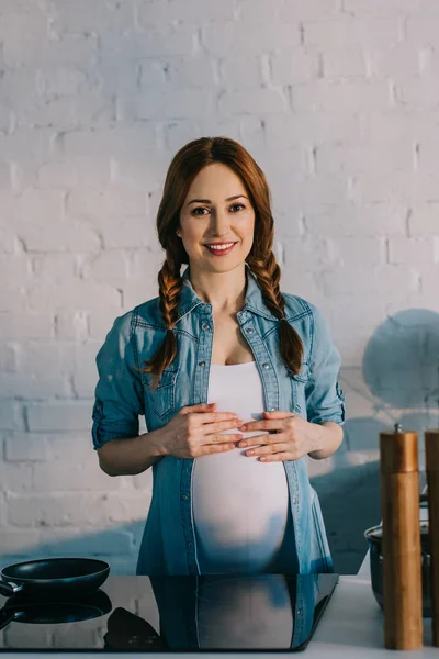 Attraente donna incinta in piedi vicino stufa elettrica in cucina — Foto stock
