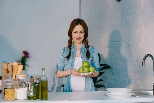 Attractive pregnant woman holding plate with ripe apples at kitchen — Stock Photo