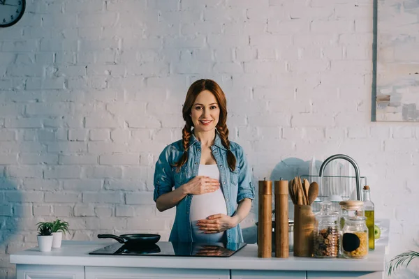 Mulher grávida atraente tocando barriga perto do fogão elétrico na cozinha — Fotografia de Stock