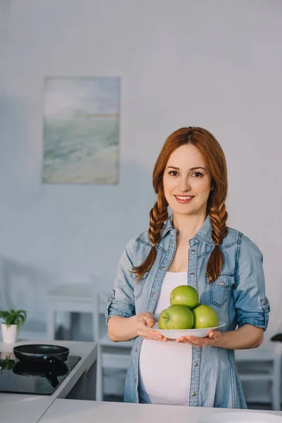Hermosa mujer embarazada sosteniendo placa con manzanas maduras en la cocina - foto de stock
