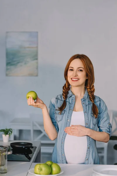Улыбающаяся беременная женщина показывает яблоко на кухне — стоковое фото