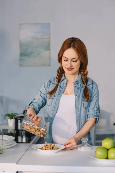 Atractiva mujer embarazada vertiendo copos de maíz en la placa en la cocina - foto de stock