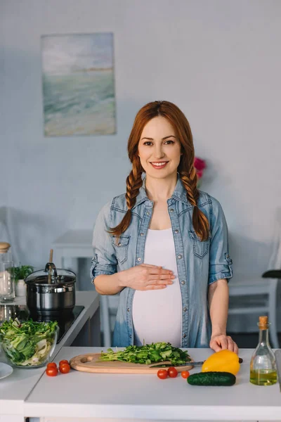 Atractiva mujer embarazada tocando el vientre cerca del mostrador de cocina con verduras para ensalada - foto de stock