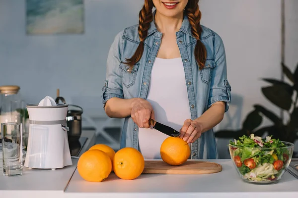 Cropped image of smiling pregnant woman cutting oranges at kitchen — Stock Photo