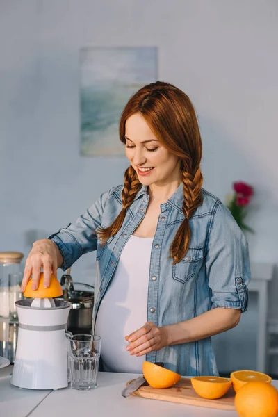 Attractive pregnant woman preparing homemade orange juice at kitchen — Stock Photo