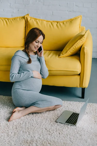 Привлекательная беременная женщина сидит на полу и разговаривает со смартфоном в гостиной — стоковое фото