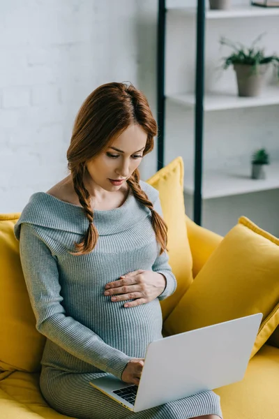 Atractiva mujer embarazada tocando el vientre y el uso de ordenador portátil en la sala de estar - foto de stock