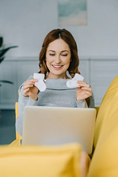 Happy pregnant woman showing newborn shoes during video call in living room — Stock Photo