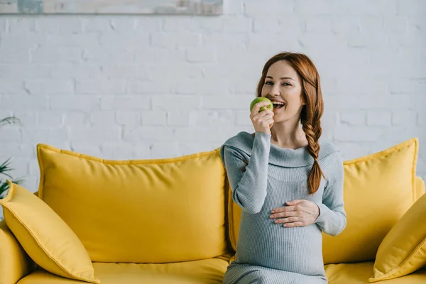 Feliz mujer embarazada atractiva comiendo manzana en la sala de estar - foto de stock