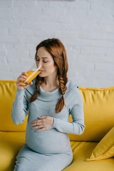 Atractiva mujer embarazada bebiendo jugo de naranja en la sala de estar - foto de stock