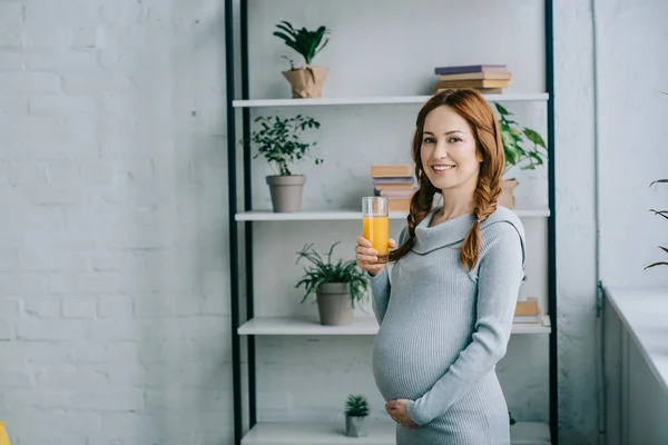 Attraente donna incinta sorridente in possesso di un bicchiere di succo d'arancia e guardando la fotocamera a casa — Foto stock