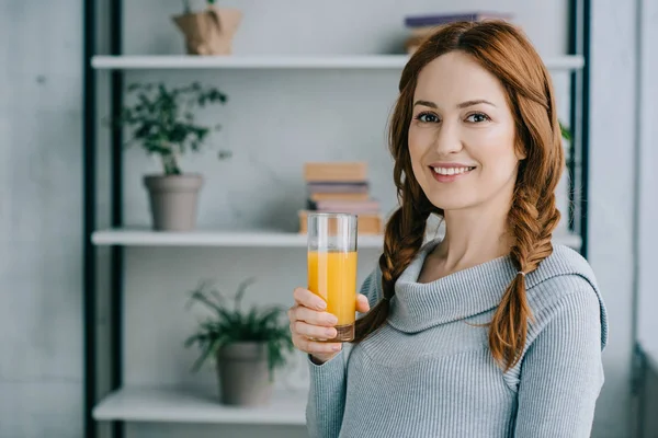 Mulher sorridente atraente segurando vidro de suco de laranja e olhando para a câmera em casa — Fotografia de Stock