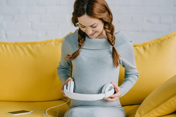 Hermosa mujer embarazada poner auriculares con música en el vientre en la sala de estar - foto de stock
