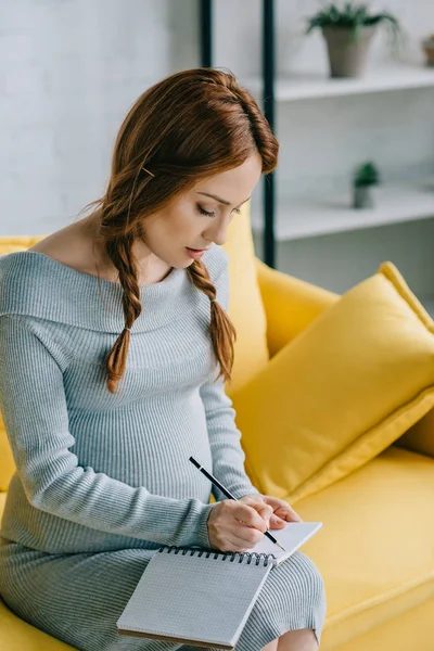 Atractiva mujer embarazada escribiendo algo a cuaderno en la sala de estar - foto de stock