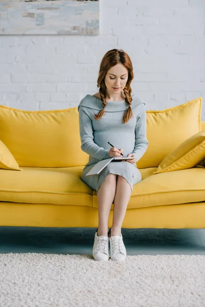 Beautiful pregnant woman sitting on sofa and writing something to notebook in living room — Stock Photo