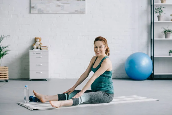 Schöne schwangere Frau dehnt sich auf Yogamatte und schaut in die Kamera im Wohnzimmer — Stockfoto