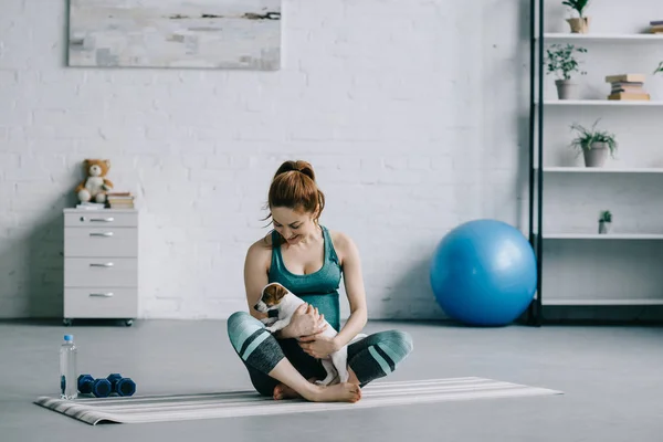 Hermosa mujer embarazada sentado en yoga mat con jack russell terrier cachorro - foto de stock