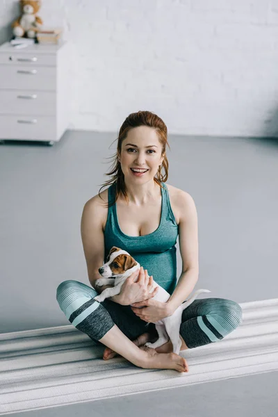 Beautiful pregnant woman sitting on yoga mat with puppy — Stock Photo