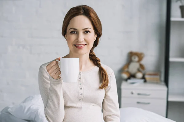 Sonriente hermosa mujer embarazada sosteniendo taza de té y mirando a la cámara en el dormitorio - foto de stock