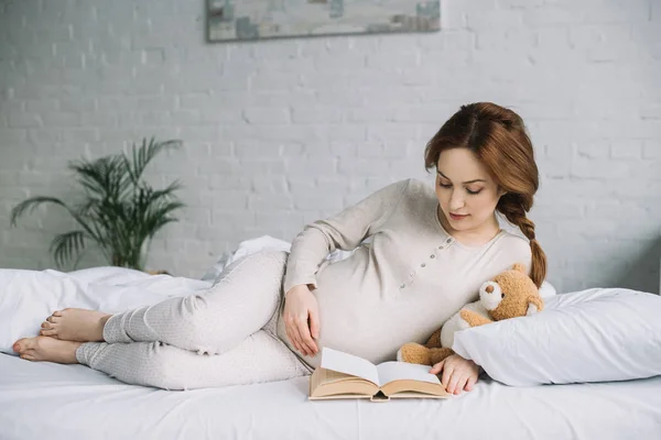 Attractive pregnant woman lying on bed with teddy and reading book in bedroom — Stock Photo
