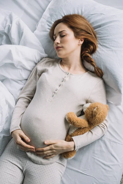 Vista de ángulo alto de hermosa mujer embarazada durmiendo en la cama con oso de peluche - foto de stock