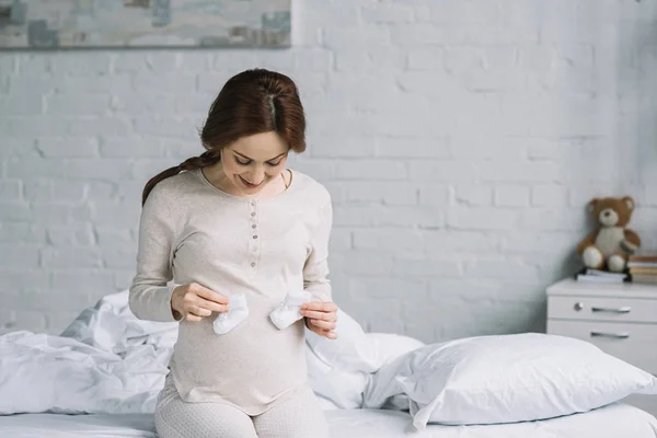 Beautiful pregnant woman touching belly with newborn shoes in bedroom — Stock Photo