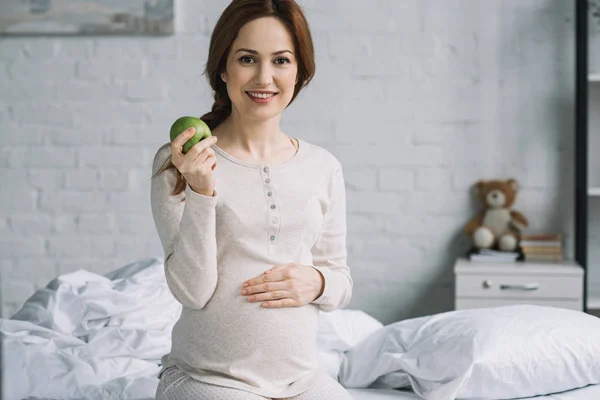 Beautiful pregnant woman sitting on bed with ripe apple — Stock Photo