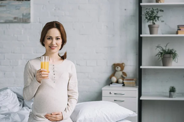 Beautiful pregnant woman sitting on bed in bedroom with glass of orange juice — Stock Photo