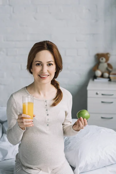 Donna incinta sorridente che tiene un bicchiere di succo d'arancia e mela in camera da letto — Foto stock