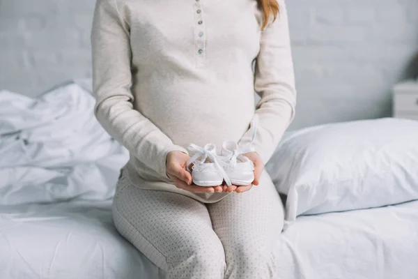Abgeschnittenes Bild einer schwangeren Frau mit weißen kleinen neugeborenen Schuhen im Schlafzimmer — Stockfoto