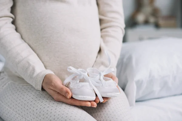 Imagen recortada de mujer embarazada sosteniendo zapatos recién nacidos en el dormitorio - foto de stock