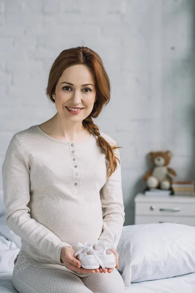 Beautiful pregnant woman holding newborn shoes and looking at camera in bedroom — Stock Photo
