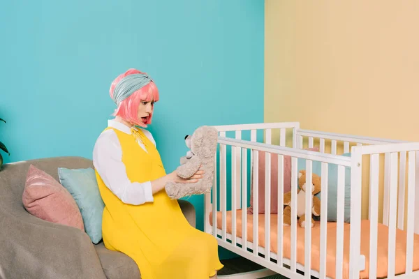 Sorprendido retro estilo embarazada pin up mujer con pelo rosa sentado con oso de peluche cerca de cuna en habitación de niño - foto de stock